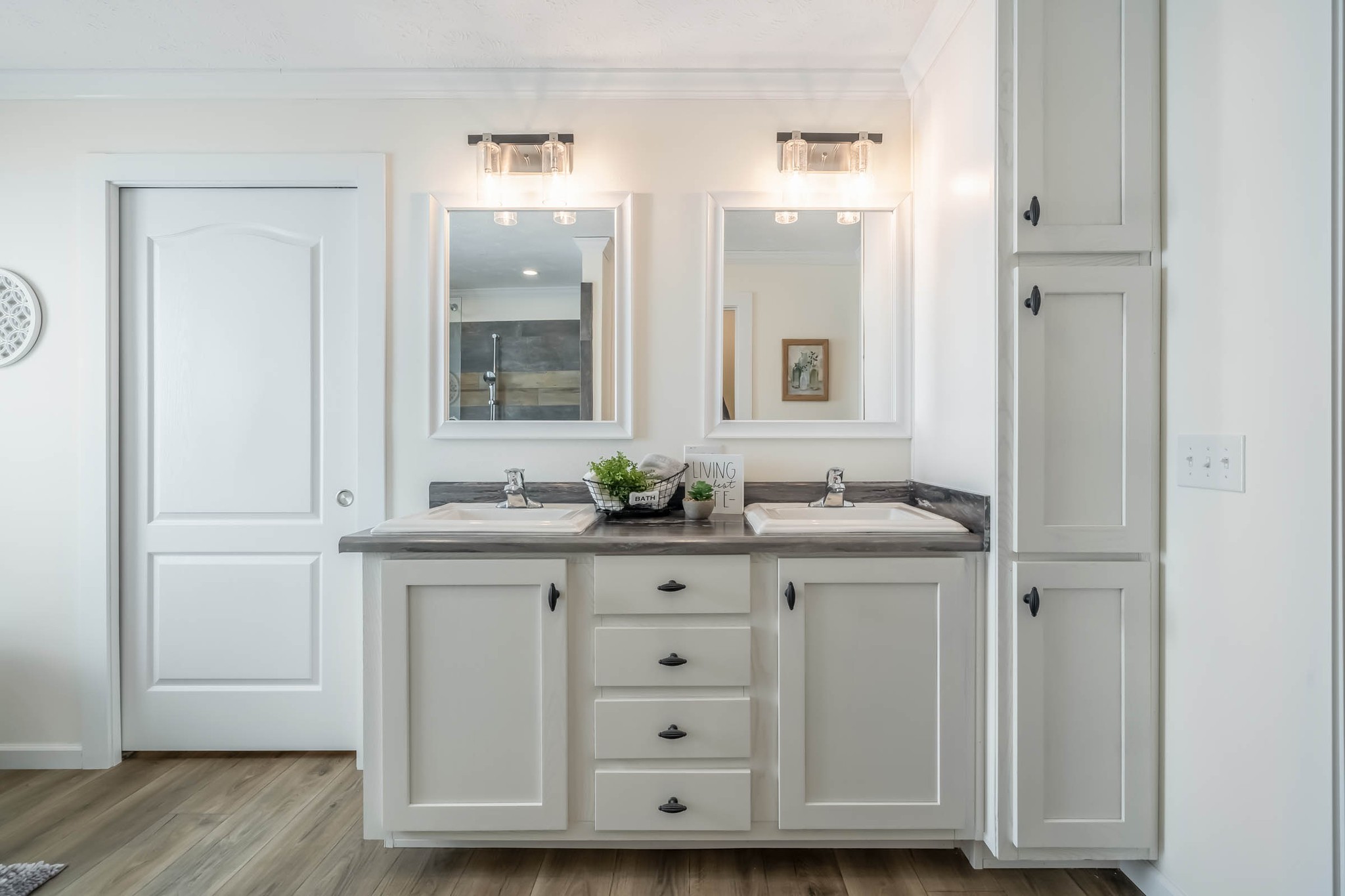 A modern bathroom features a double sink vanity with white cabinets, dark countertops, and two mirrors. Soft lighting and minimal decor create a clean, calming ambiance.