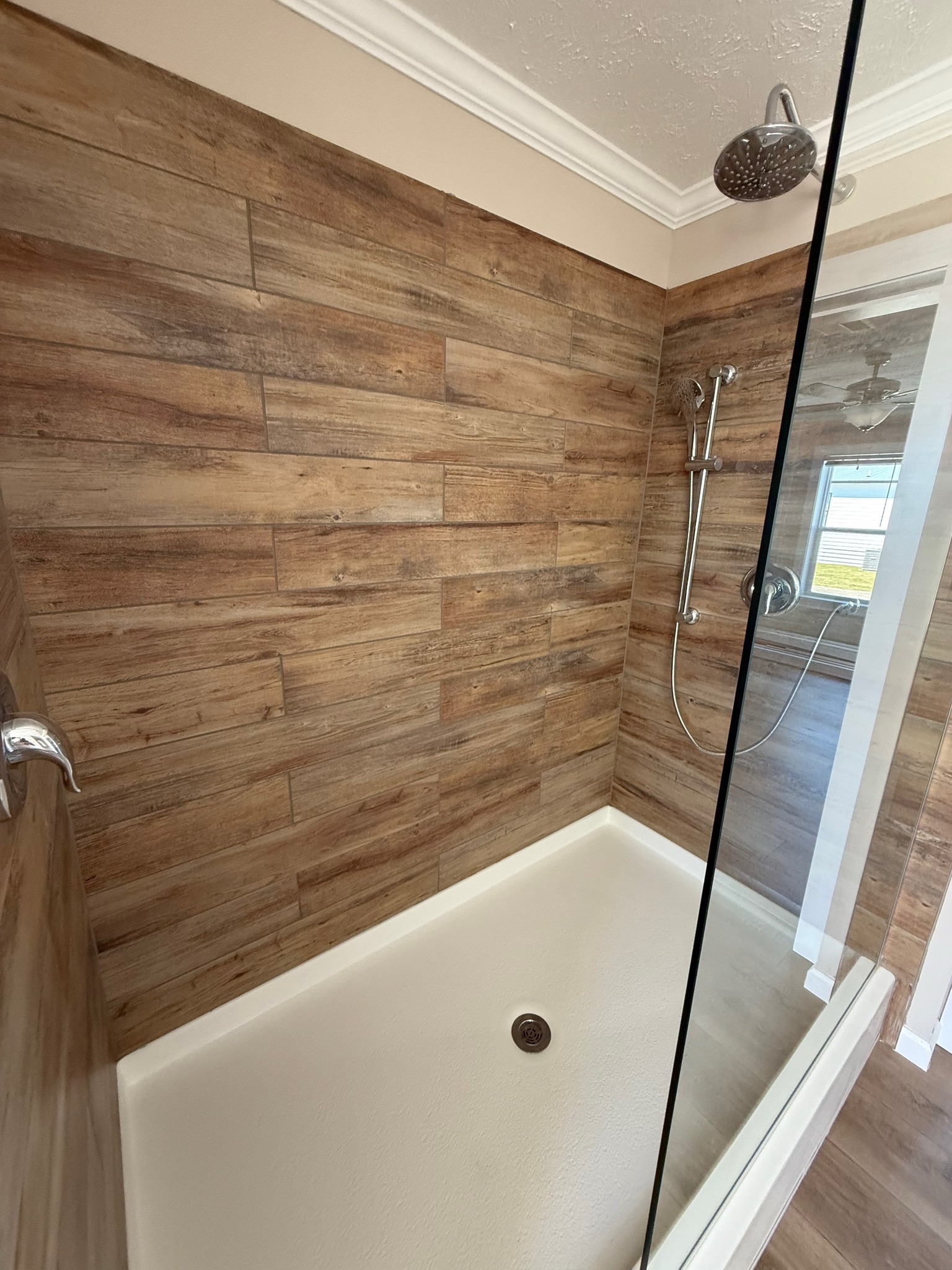 Modern shower with wood-style tiled walls, a large shower head, handheld showerhead, glass door, and white base. Bright, clean, and minimalist tone.