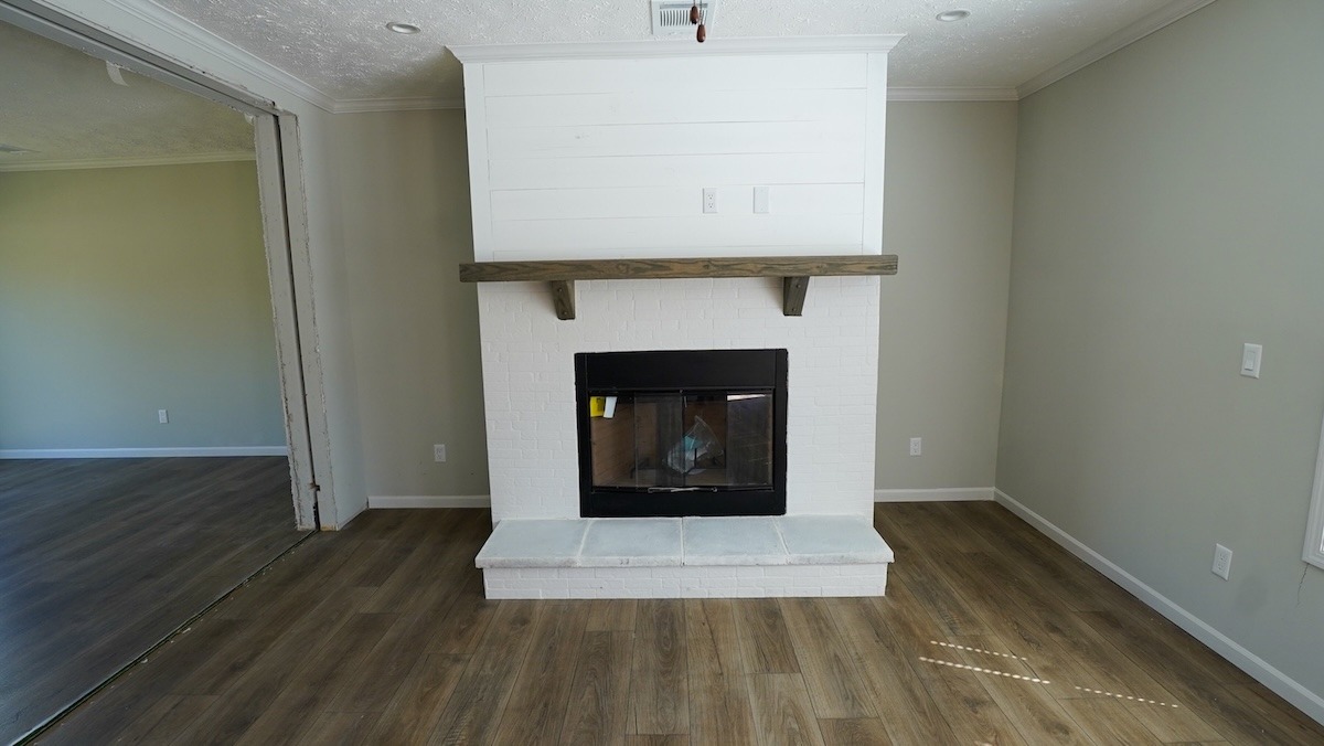 Room with wood floors, a white brick fireplace topped by a wooden mantel. Adjacent room visible through a large opening; neutral walls, bright atmosphere.