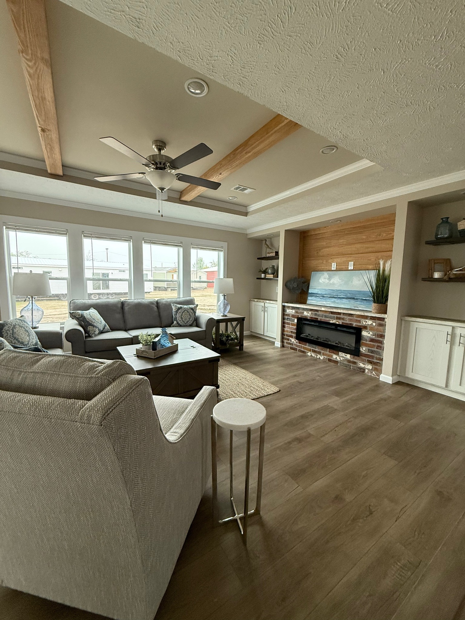 Stylish living room with gray sofas, brick fireplace, and large windows. Ceiling features wood beams and fan. Neutral tones, cozy atmosphere.