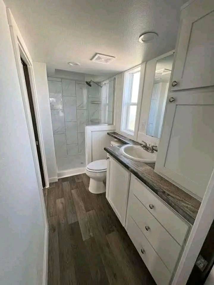 Narrow bathroom with wood floor, featuring a glass shower, white toilet, and a counter with a sink and cabinets beneath. Bright and minimalist.