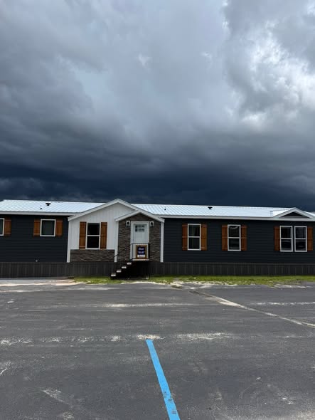 A dark gray modular building with wooden shutters and a metal roof sits under a stormy sky, conveying a tense and ominous atmosphere.
