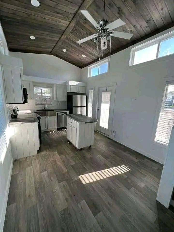 Modern kitchen with a dark wooden ceiling and fan, light wood flooring, white cabinets, and stainless steel appliances. Sunlight streams through high windows.