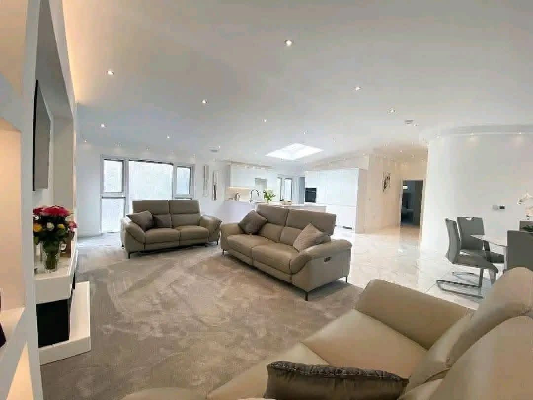 Spacious modern living room with gray sofas, soft carpet, and a skylight. Open kitchen and dining area with sleek finishes and fresh flowers add elegance.
