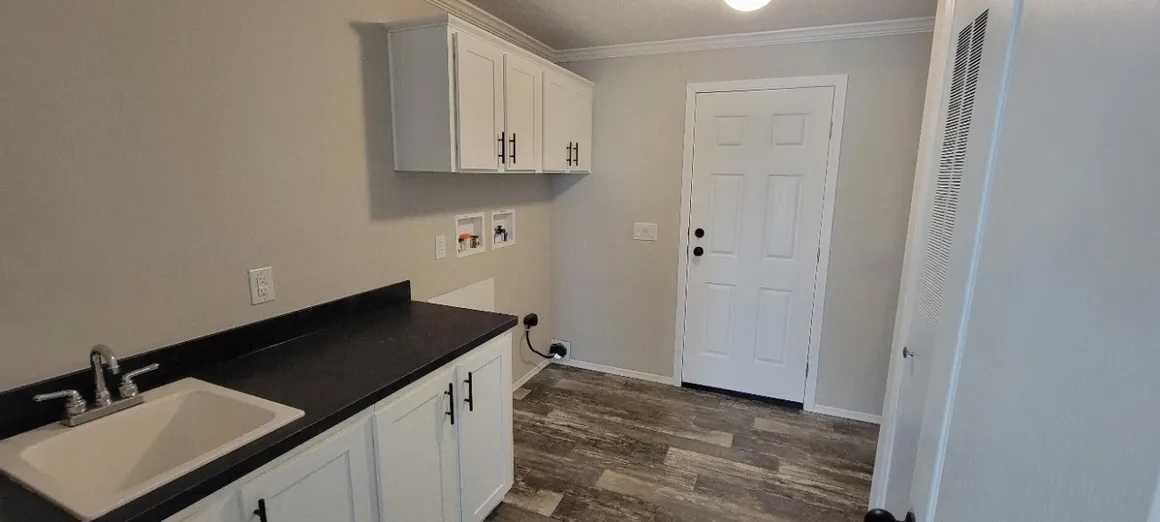 A clean laundry room with gray flooring. It features white cabinets, a black countertop, a sink, and a white door. The space is neat and minimalistic.