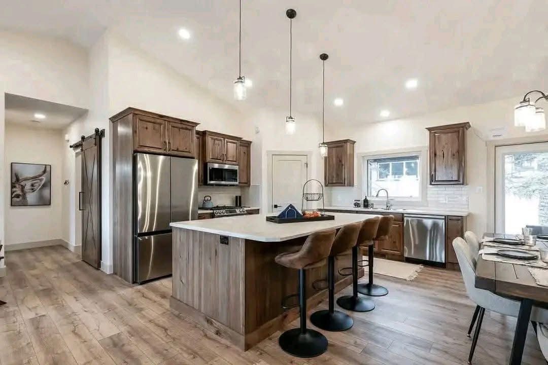 Spacious kitchen with wooden cabinets, center island with brown leather stools, stainless steel appliances. Cozy dining table to the right, bright lighting.