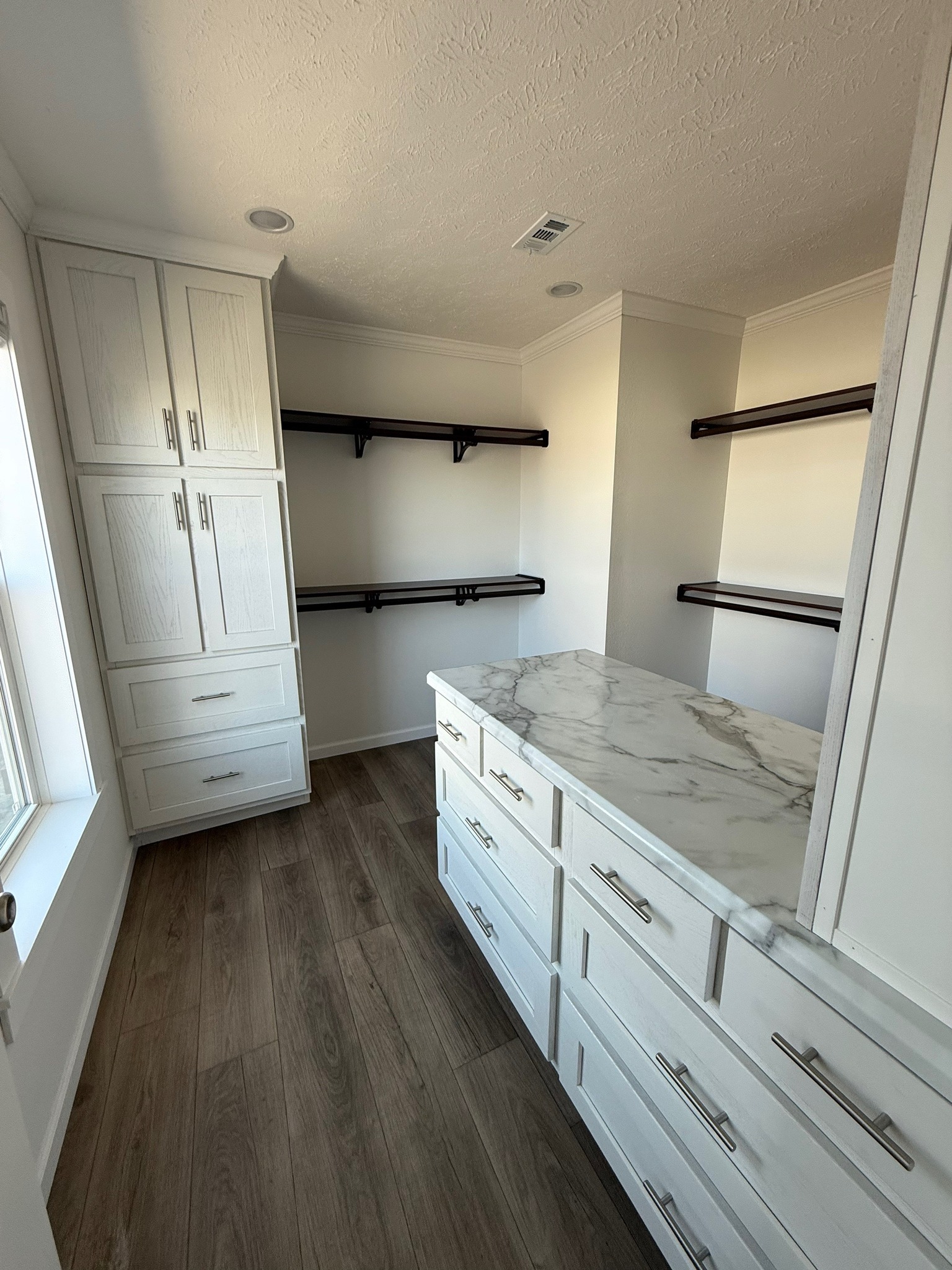 A modern walk-in closet featuring a white marble-topped island with drawers, white cabinets, dark wood shelves, and light wood flooring, creating a clean and organized look.