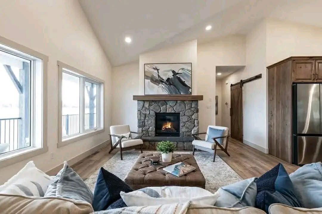 Cozy living room with stone fireplace, abstract art, and modern chairs. Large windows, cushioned ottoman, light wood floors, inviting atmosphere.