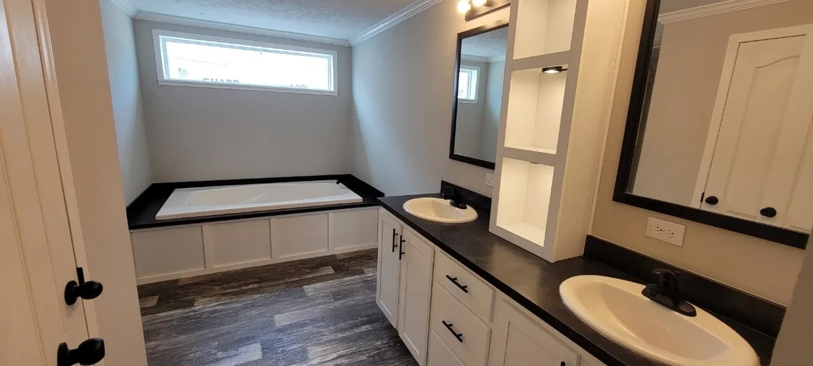 Modern bathroom with a large, rectangular bathtub under a horizontal window. Double sinks on a black countertop, white cabinetry, and wood-like flooring.