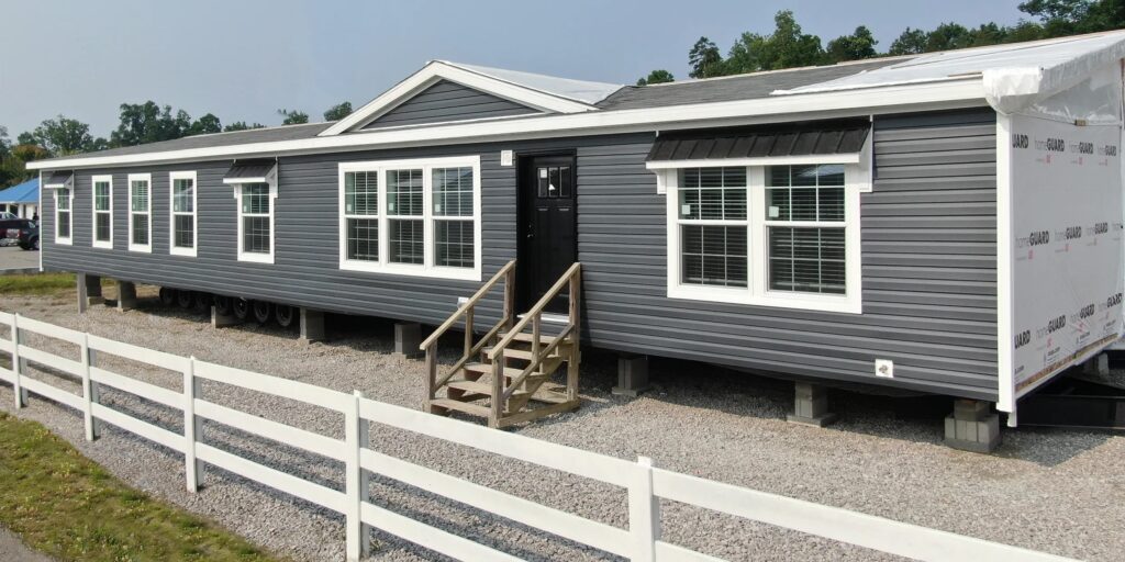 A gray modular home with white-trimmed windows and a center black door stands on a gravel lot. A white fence borders the front, with trees in the background.