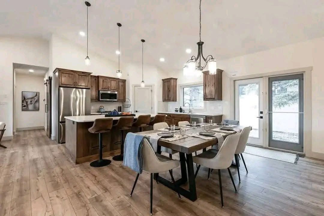 Modern kitchen-dining area with wooden floors, a large wooden table, beige chairs, pendant lights, and stainless steel appliances. Airy and inviting.