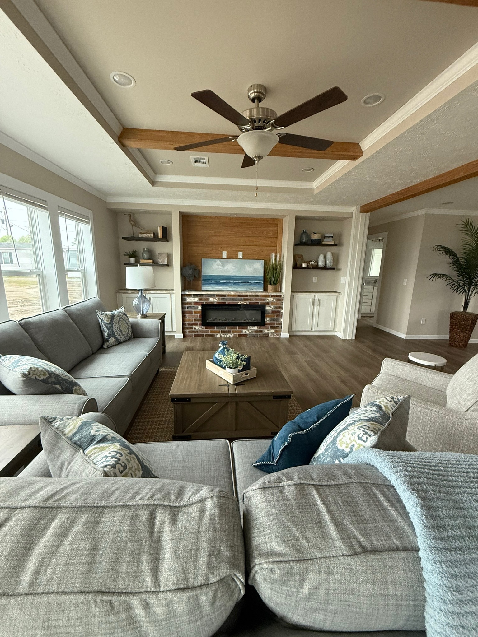 Modern living room with gray sofas, blue and patterned cushions, a wooden coffee table, and a TV above a brick fireplace. Ceiling fan and wood beams add warmth.