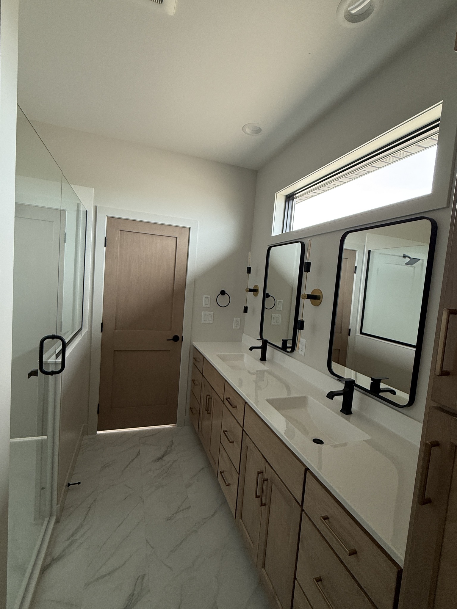 Modern bathroom with light wood cabinets, dual sinks, and black framed mirrors. A wooden door and glass-enclosed shower add a clean, minimalist feel.
