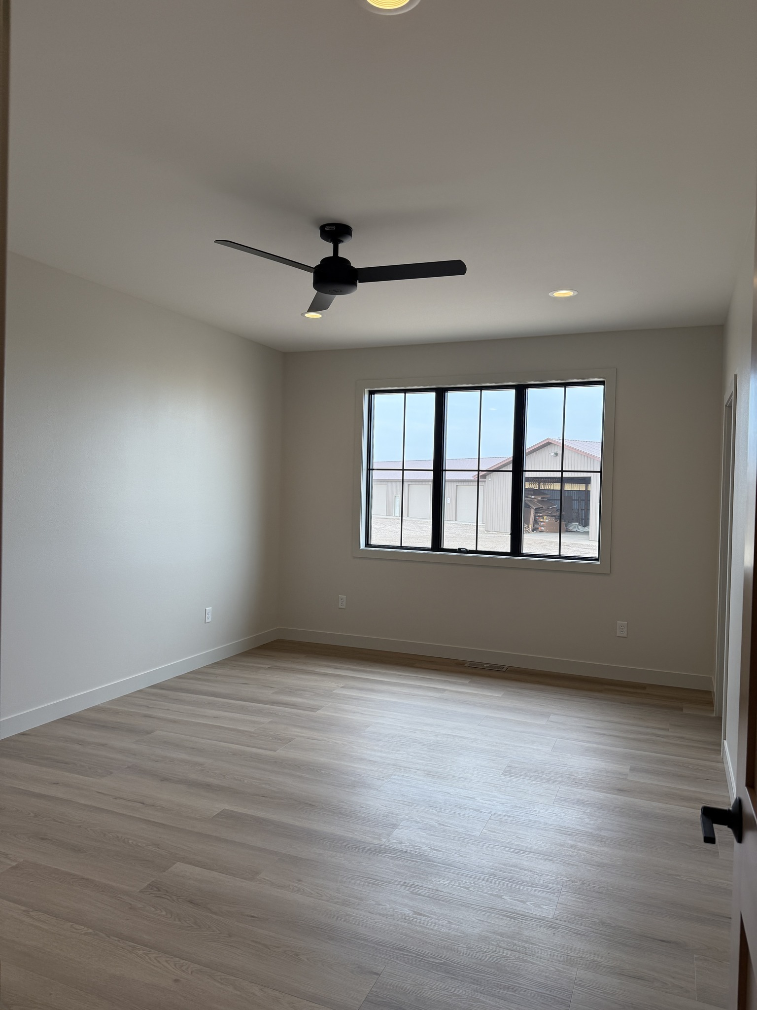 Empty room with light wood flooring, white walls, and a large window showing an exterior building. Ceiling fan and recessed lights add a modern touch.