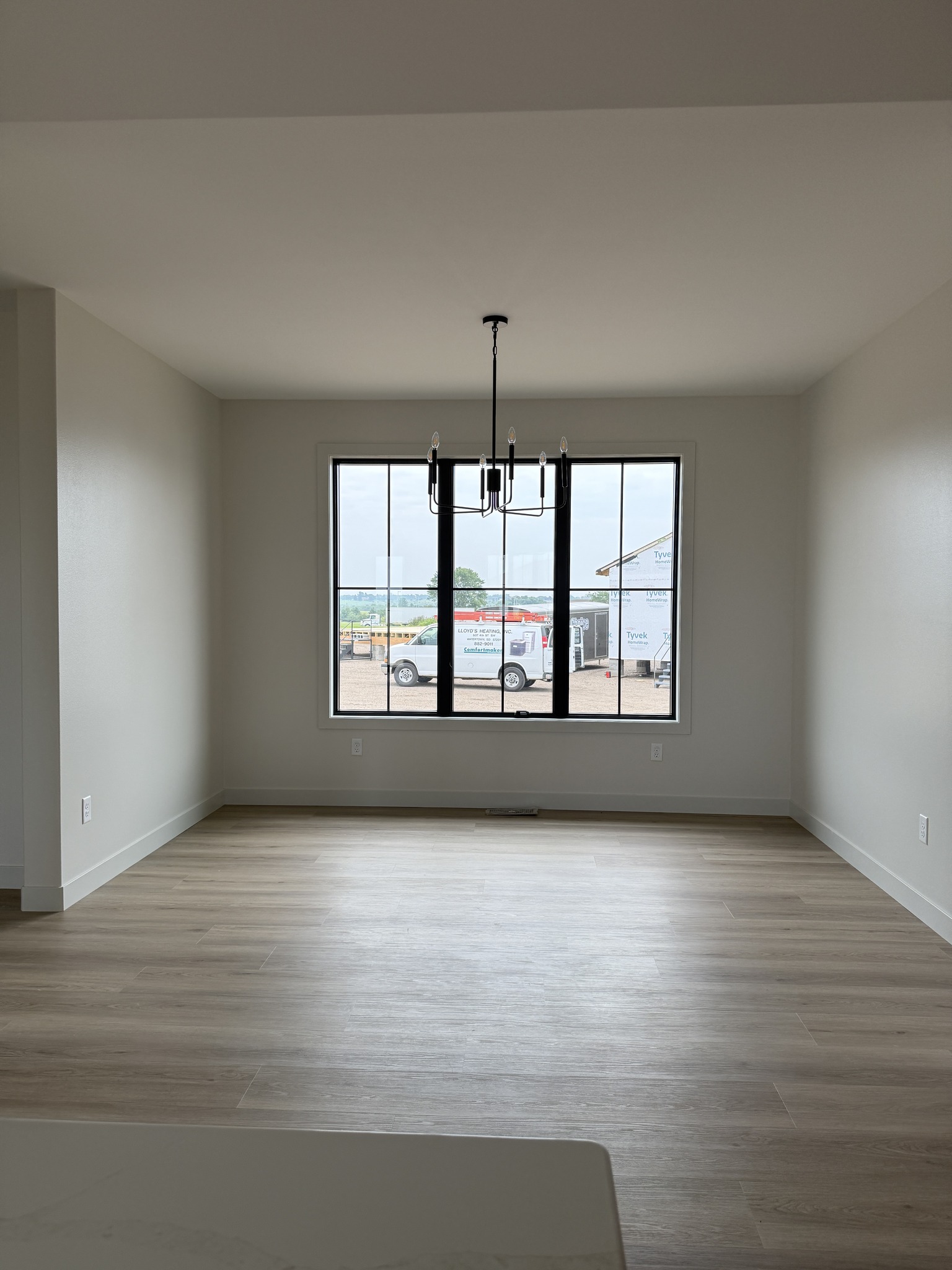 Bright empty room with light wood floor, white walls, large window showing a construction site, and a modern black chandelier, conveying openness.