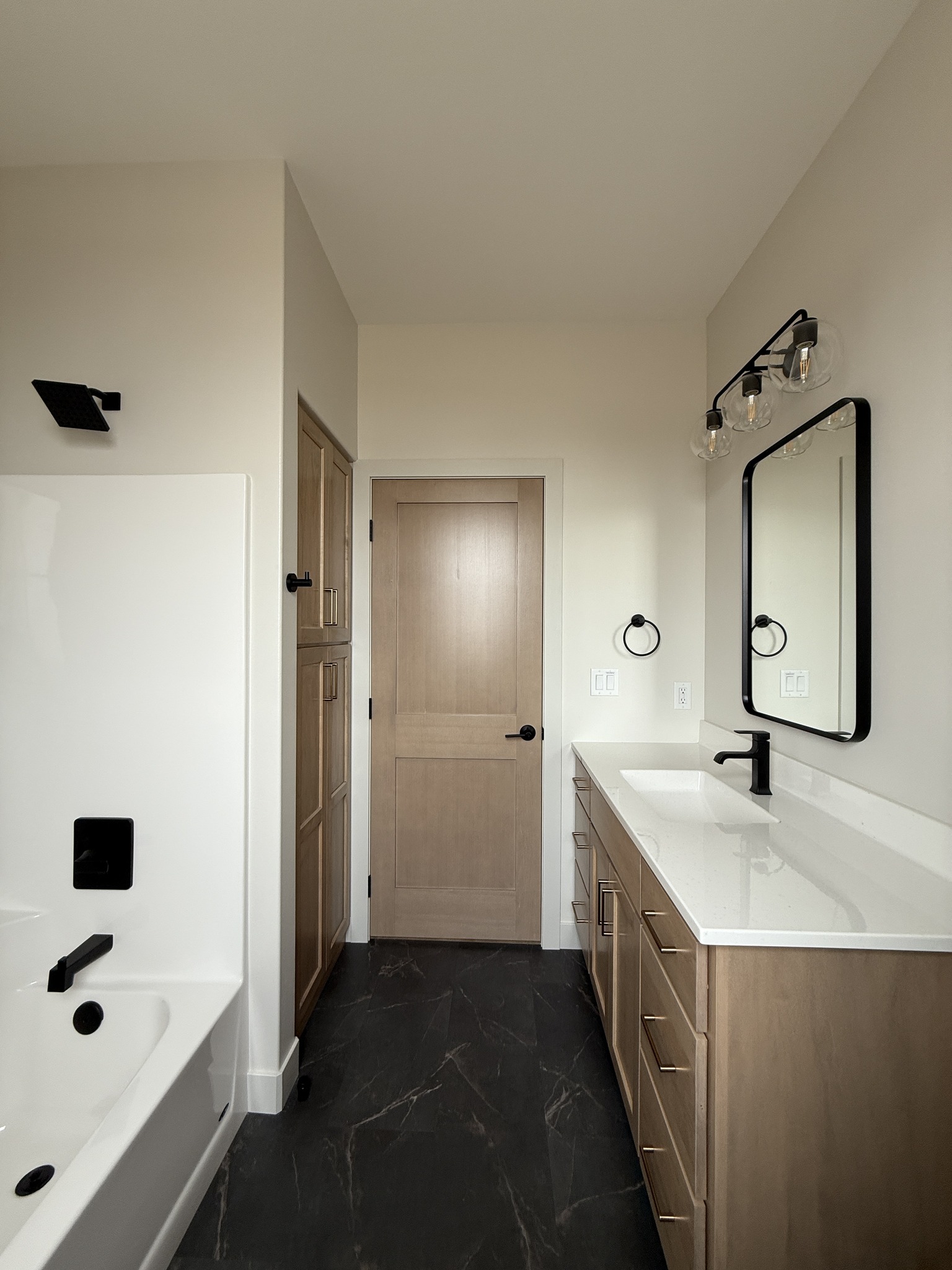 Modern bathroom with light wood cabinetry, white countertop, and large mirror. Black fixtures contrast with neutral tones. Bathtub on the left.