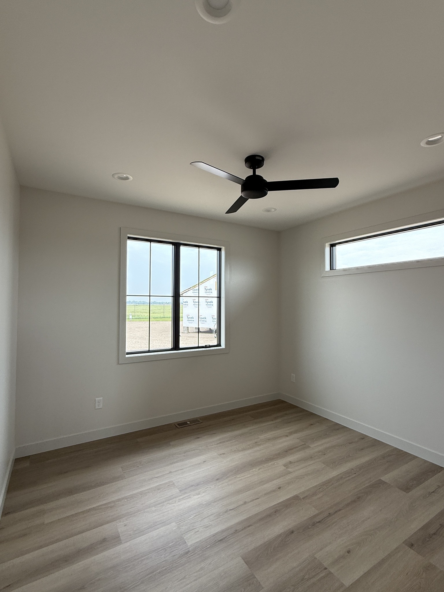 A small, empty room with light wood flooring and white walls. It has a ceiling fan and two windows, one with a view of a clear sky and grassy area.