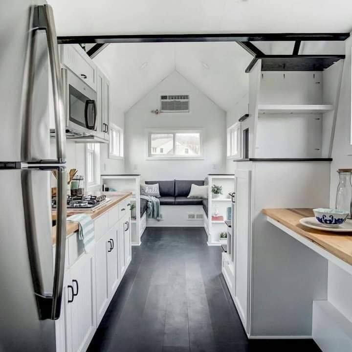 Bright, modern tiny house interior with white cabinetry, stainless steel appliances, and a cozy seating area in the back. Airy, minimalist design.