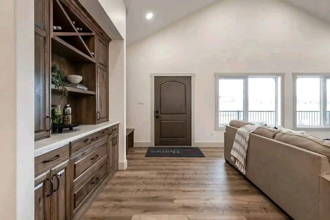 Spacious living room with wooden floors and light walls. A large brown cabinet with decor on the left, beige couch on the right. Door and windows in the background.