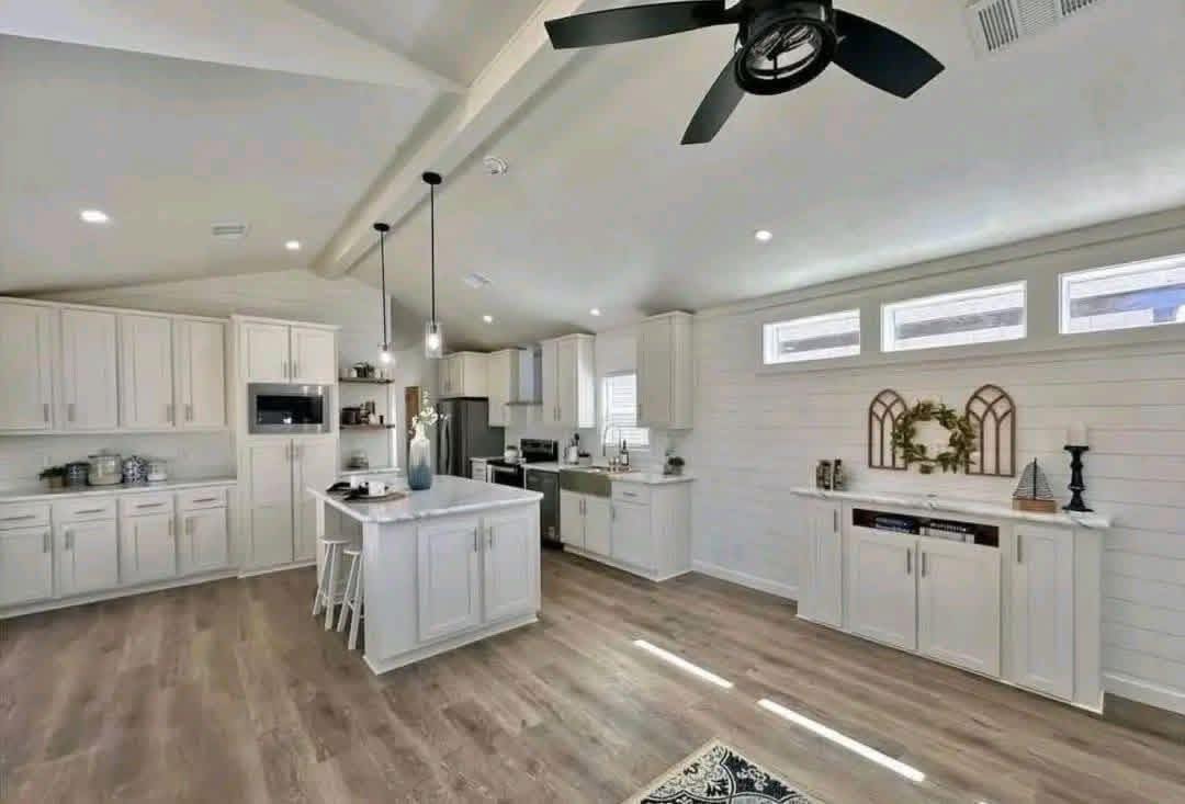Spacious modern kitchen with white cabinets, a central island with stools, pendant lighting, wood flooring, and a ceiling fan, creating a bright, airy feel.