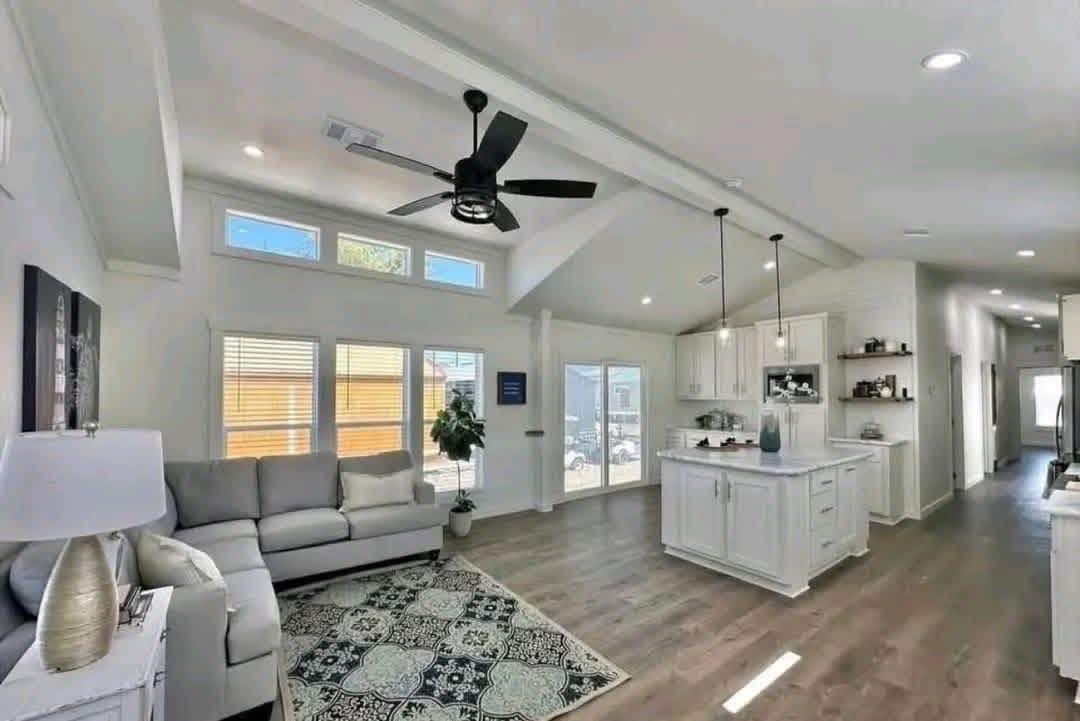 Spacious living area with a modern kitchen, featuring white cabinets, a central island, pendant lights, large windows, and a cozy gray sofa.