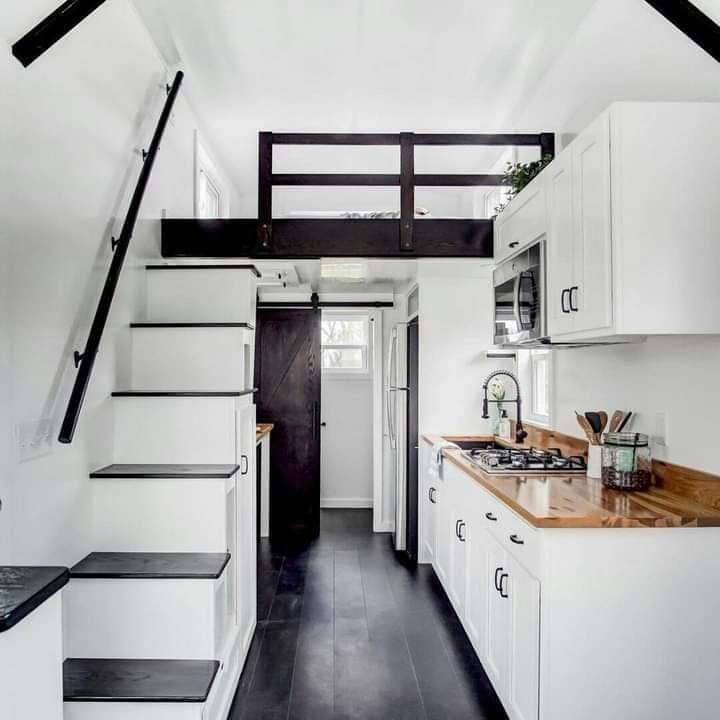 Modern tiny house interior with sleek white cabinets, wooden countertops, and a black railing leading to a loft. Cozy and minimalist vibe.