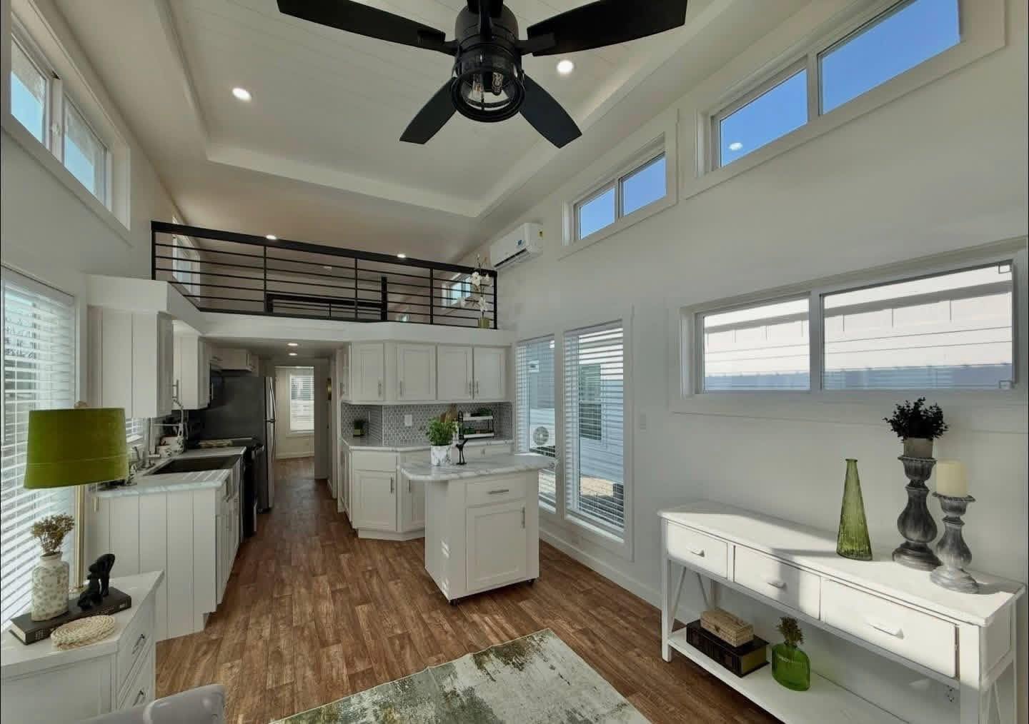 Modern tiny home interior with high ceiling, wooden floor, and white walls. Features a loft, compact kitchen, and minimal decor, creating an airy feel.