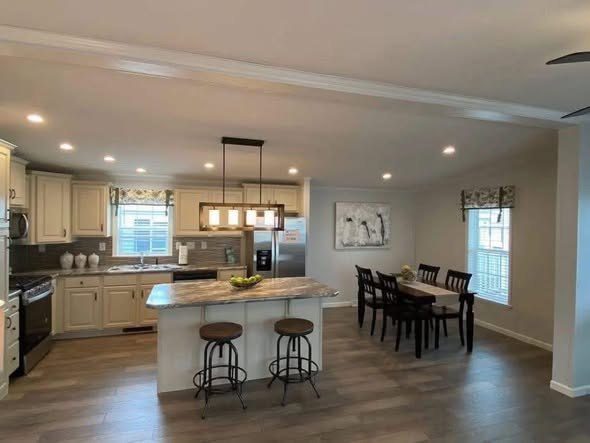 A spacious, modern kitchen and dining area with light wood flooring. Features an island with two stools, elegant lighting, and a table for six.