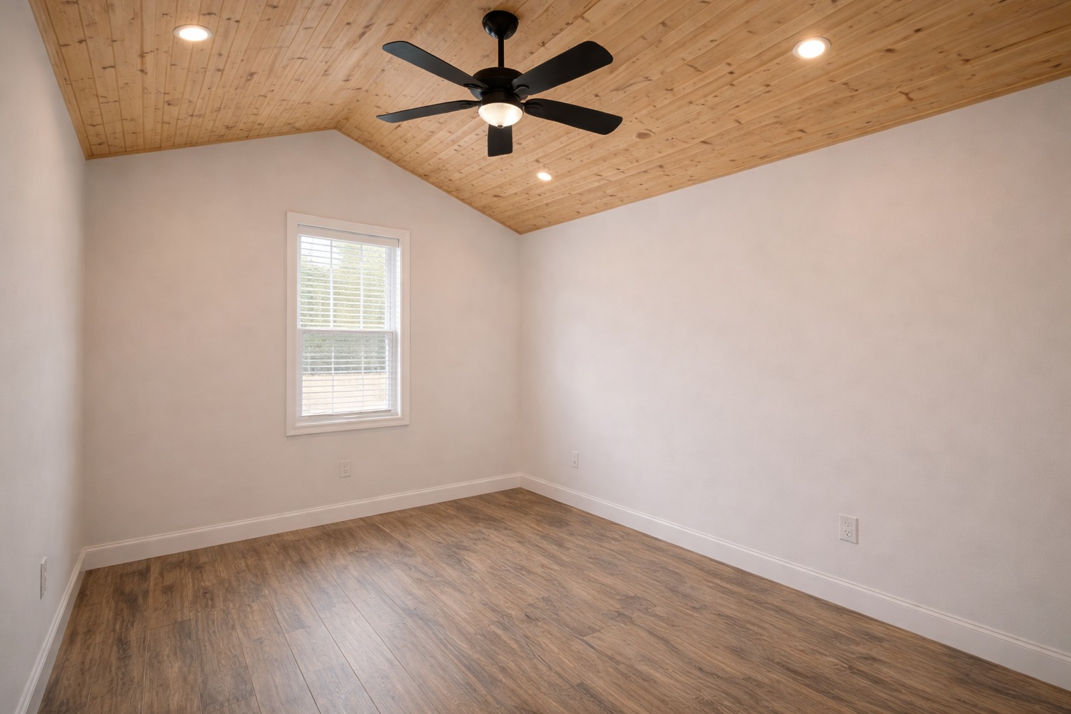 A minimalist room with wooden flooring and a matching wood-paneled ceiling featuring recessed lights. A black ceiling fan and a single window add light and contrast.
