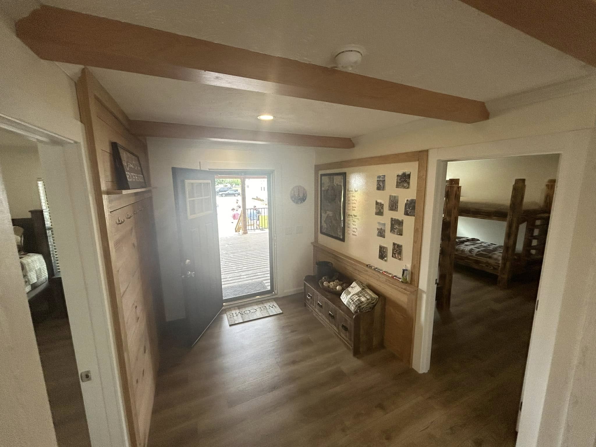Cozy entryway with wooden beams, open door leading outside, family photos on walls, wooden bench with storage, and adjoining bedrooms featuring bunk beds.