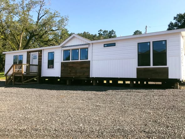 Single-story modular home with white siding and brown accents, set on a gravel lot. It has large windows and a small wooden porch, surrounded by trees.