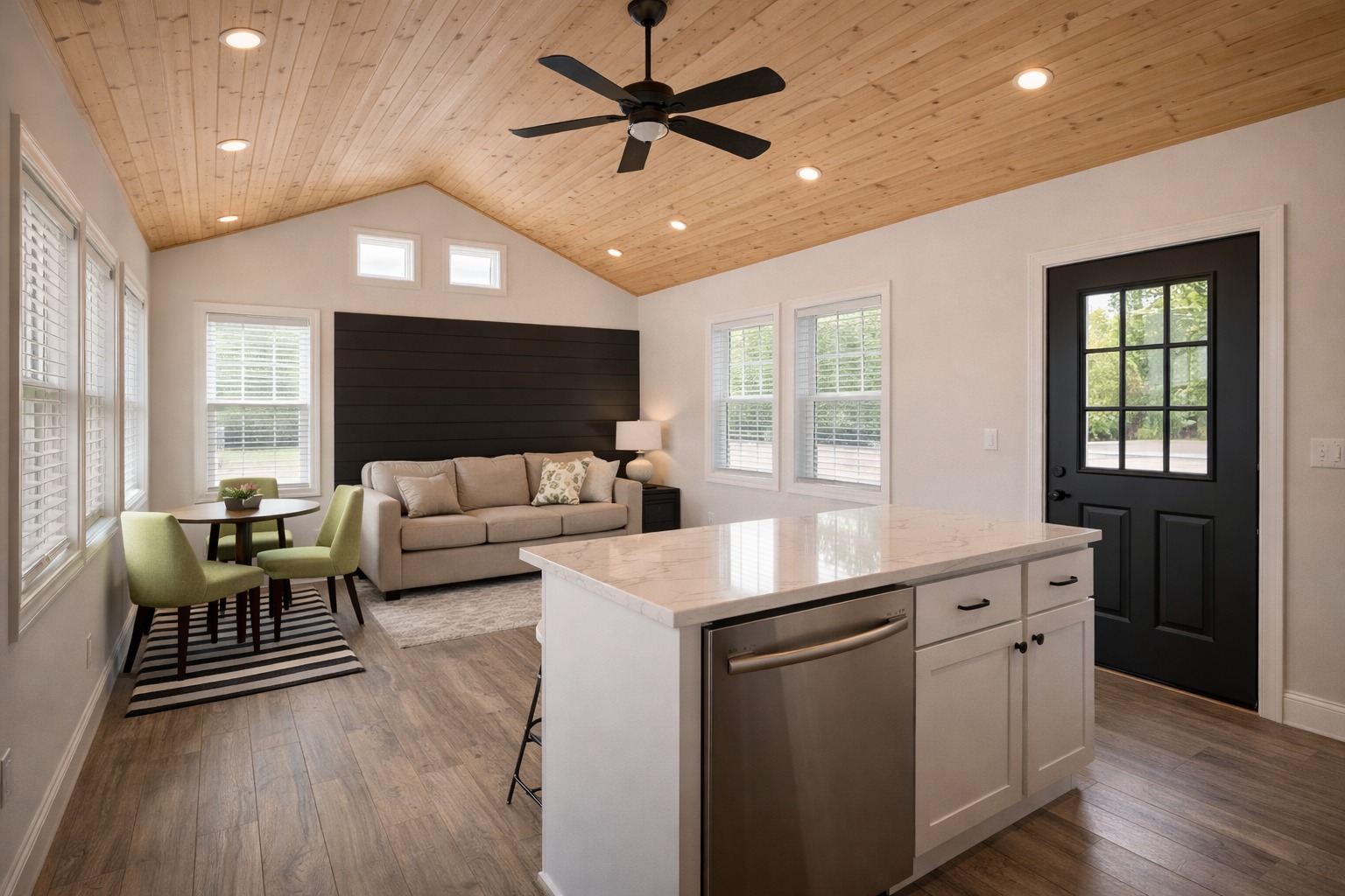 Bright living room with wooden ceiling, modern kitchen island, and cozy seating area. Features green chairs, beige sofa, and black door with glass panes.