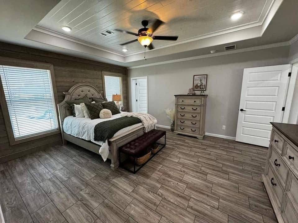 Spacious bedroom with a cozy ambiance, featuring a plush bed with green accents, wooden floors, tray ceiling, and warm lighting from a ceiling fan.