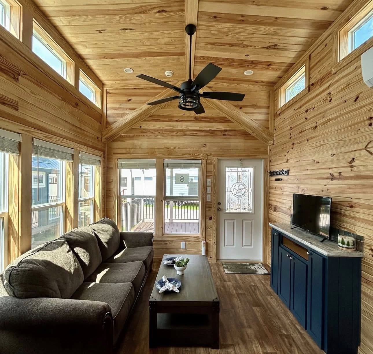 High-end housing showcase featuring a diverse collection of modern manufactured homes, tiny homes, and park models, including the expansive "The Boujee XL" aesthetic.

Exterior Architectural Styles: Highlights include classic white board-and-batten tiny homes and park models with grand pillared front porches, black metal railings, and sleek metal roofing. A premium model features a striking dark charcoal exterior with a copper-tone metal roof, natural cedar wood accents, and a large wrap-around elevated wooden deck. Also featured are traditional manufactured homes with tan vinyl siding, decorative red shutters, and spacious covered front porches.

Interior Living & Custom Design: Open-concept interiors are defined by high-fidelity finishes, including vaulted ceilings with rustic dark wood beams and white shiplap walls. Living areas showcase a "modern boujee" style with neutral-toned sofas, integrated electric fireplaces with white mantels, and lofted sleeping quarters with black metal balustrades. Traditional models offer cozy living spaces with patterned armchairs and decorative window treatments.

Gourmet Kitchens & Dining: The collection features diverse kitchen layouts, including rustic wood-cabin styles with dark marble-look countertops and high-end modern designs. Premium features include oversized kitchen islands in statement colors like teal and charcoal, white farmhouse cabinetry, recessed lighting, and elegant pendant chandeliers over dining areas with dark wood-look flooring. Professional-grade stainless steel appliances are integrated throughout.

Spa-Inspired Bathrooms & Bedrooms: Bathrooms emphasize modern luxury with walk-in tile showers featuring black matte hardware, marble-look surrounds, and contemporary vanities with ample storage. Bedrooms offer clean, minimalist designs with neutral carpeting and large windows. This gallery represents the pinnacle of "boujee" manufactured living, combining versatile floor plans with sophisticated, modern farmhouse aesthetics.