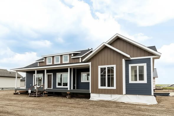 A modern single-story house with brown and blue siding sits on a dirt lot. It has a covered porch, large windows, and a cloudy sky overhead.