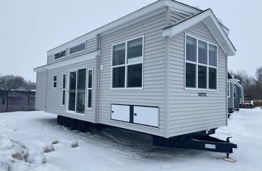 A modern tiny home on a snowy lot, with light gray siding and large windows. The overcast sky and barren trees suggest a cold, serene atmosphere.