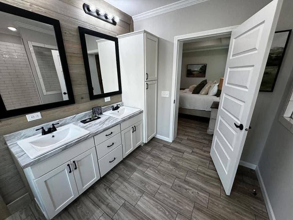 Modern bathroom with a double sink vanity, framed mirrors, and wooden accents. Open door leads to a cozy bedroom. Neutral tones create a calming atmosphere.