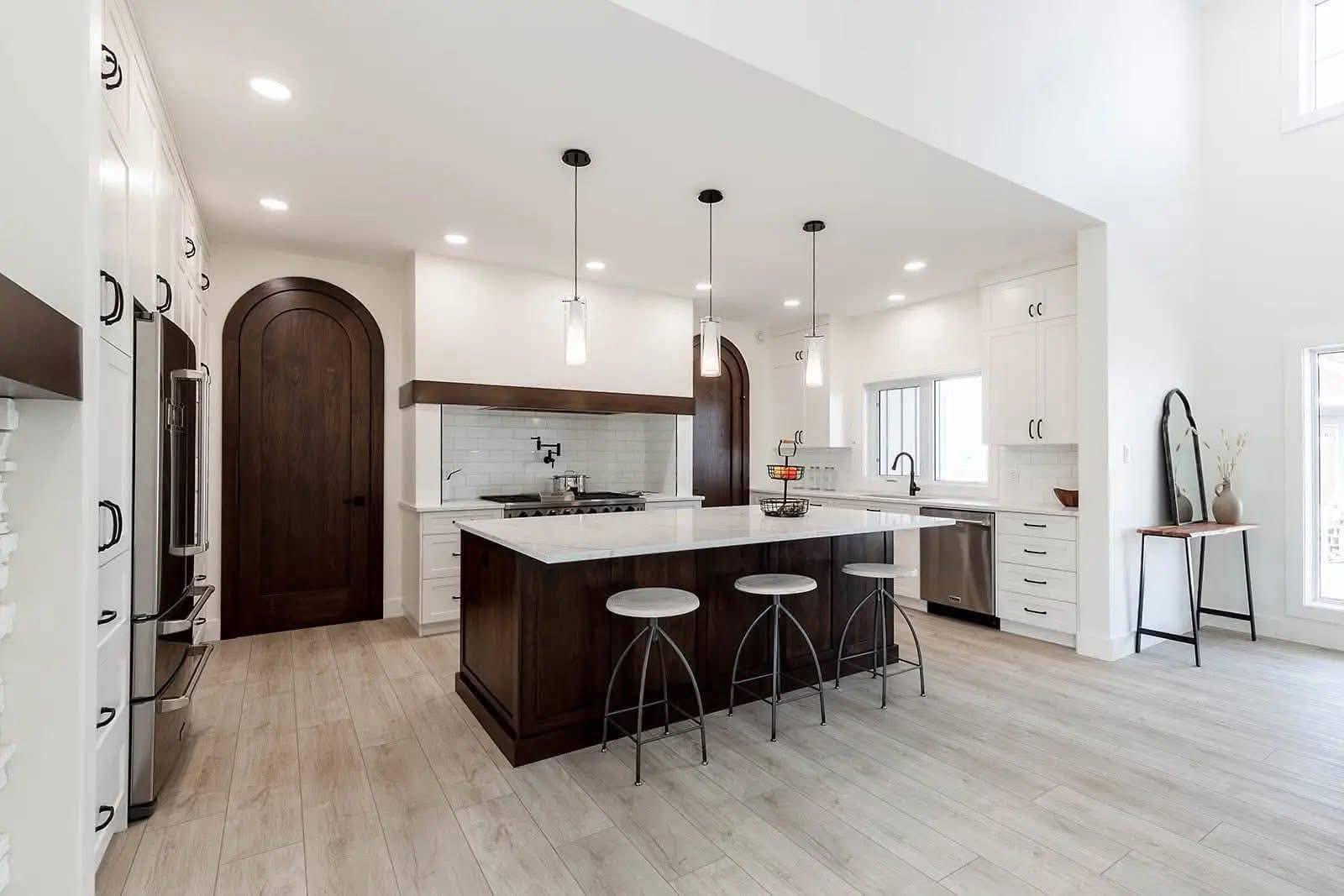 Modern kitchen with light wood floors, dark wooden island, three metal stools, pendant lights, and arched wooden doors. White cabinets and appliances.