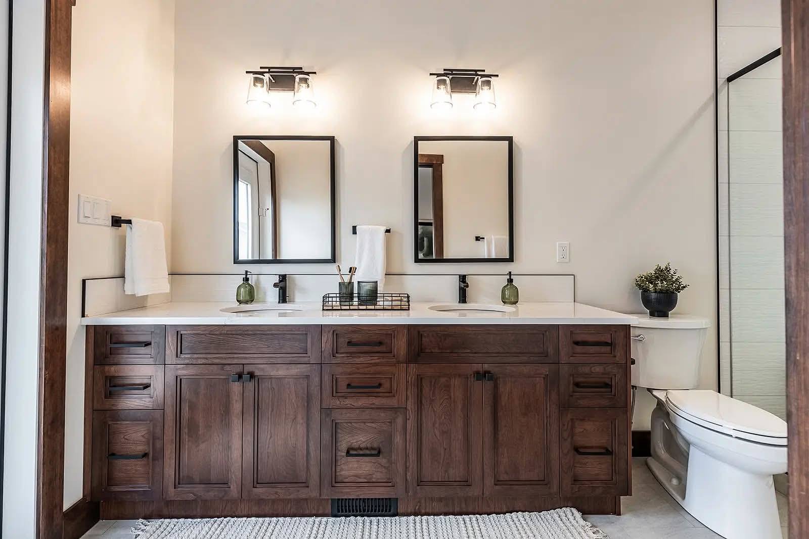 A modern bathroom features a wooden double vanity with dual sinks and mirrors. Soft lighting above and a potted plant near the toilet add a calming touch.