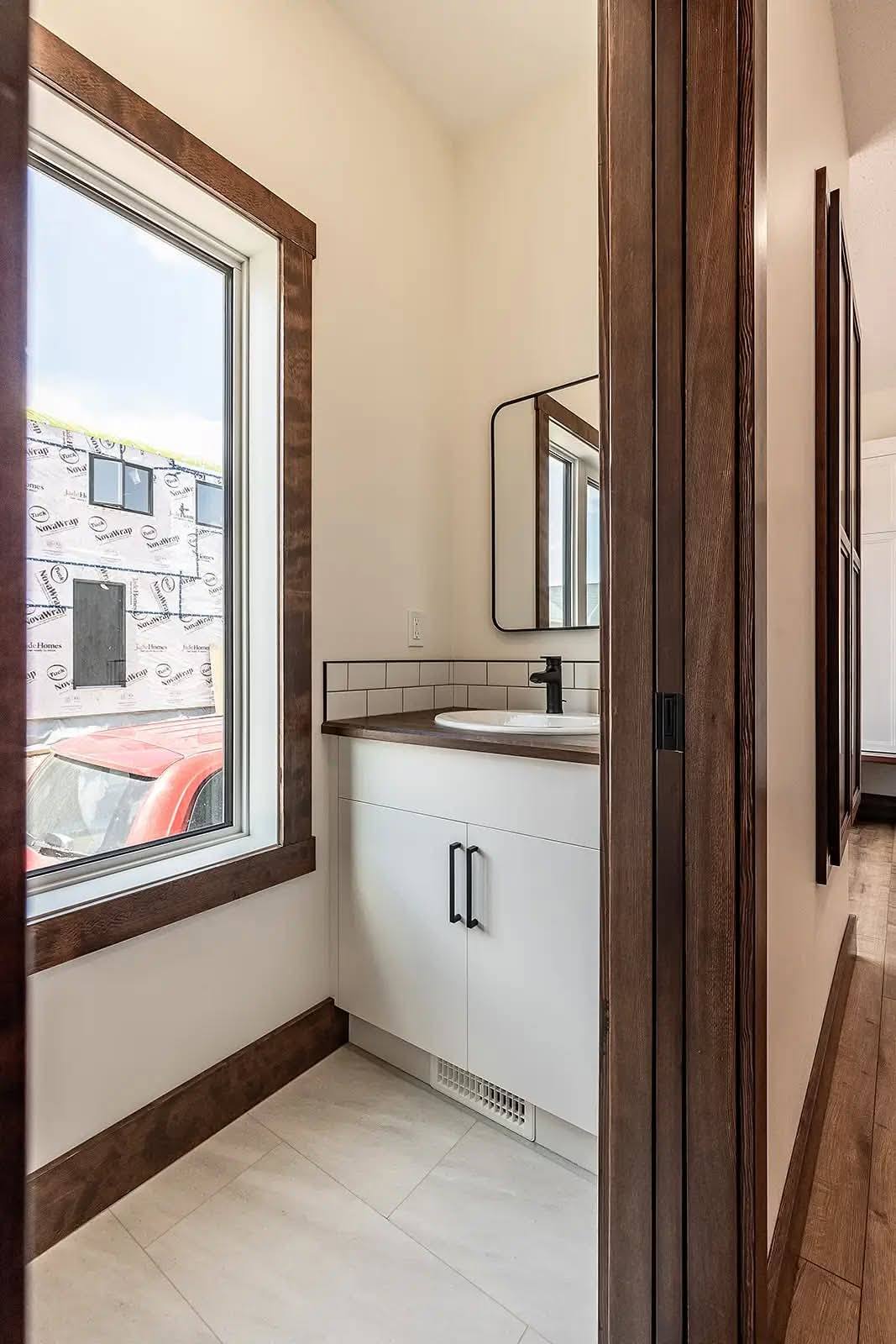Small, modern bathroom with white cabinet and black faucet. Large window with wooden trim, and a rectangular mirror above the sink. Bright and minimalistic.