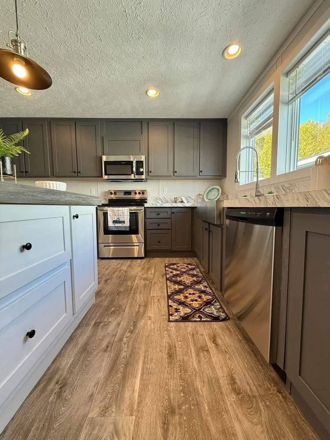 Modern kitchen with gray cabinets, stainless steel appliances, and wood flooring. Sunlight streams through large windows, creating a warm, inviting atmosphere.