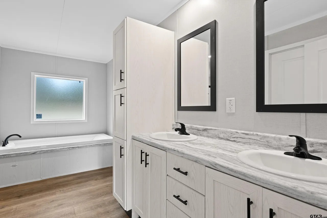 Modern bathroom with dual sinks on a marble countertop, black faucets, and framed mirrors. Light wood cabinets contrast with dark handles. Bathtub near a frosted window.