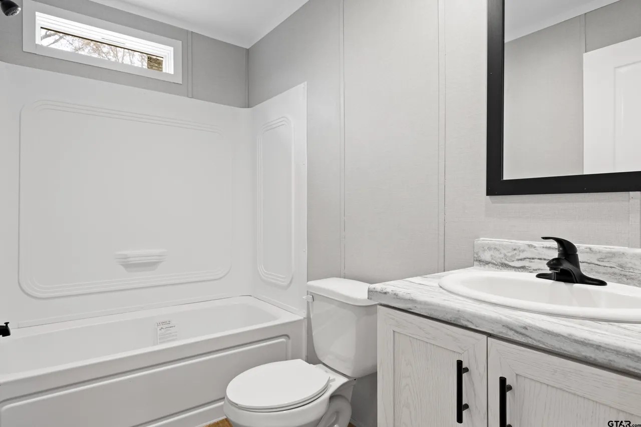 A modern bathroom with white walls features a bathtub, toilet, and sink. The countertop is marble, and a black-framed mirror hangs above.