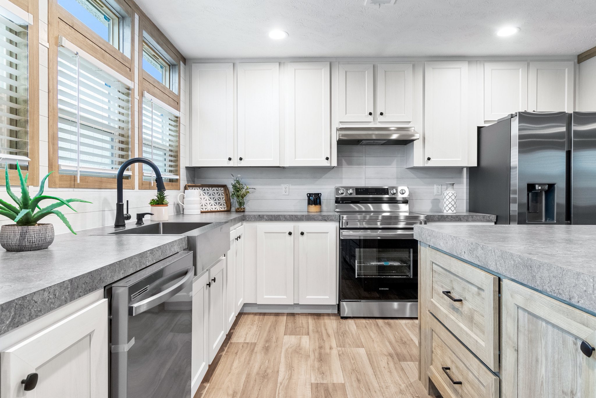 Modern kitchen with white cabinets, gray countertops, stainless steel appliances, and a double-door fridge. Light wood floors and large windows add brightness.
