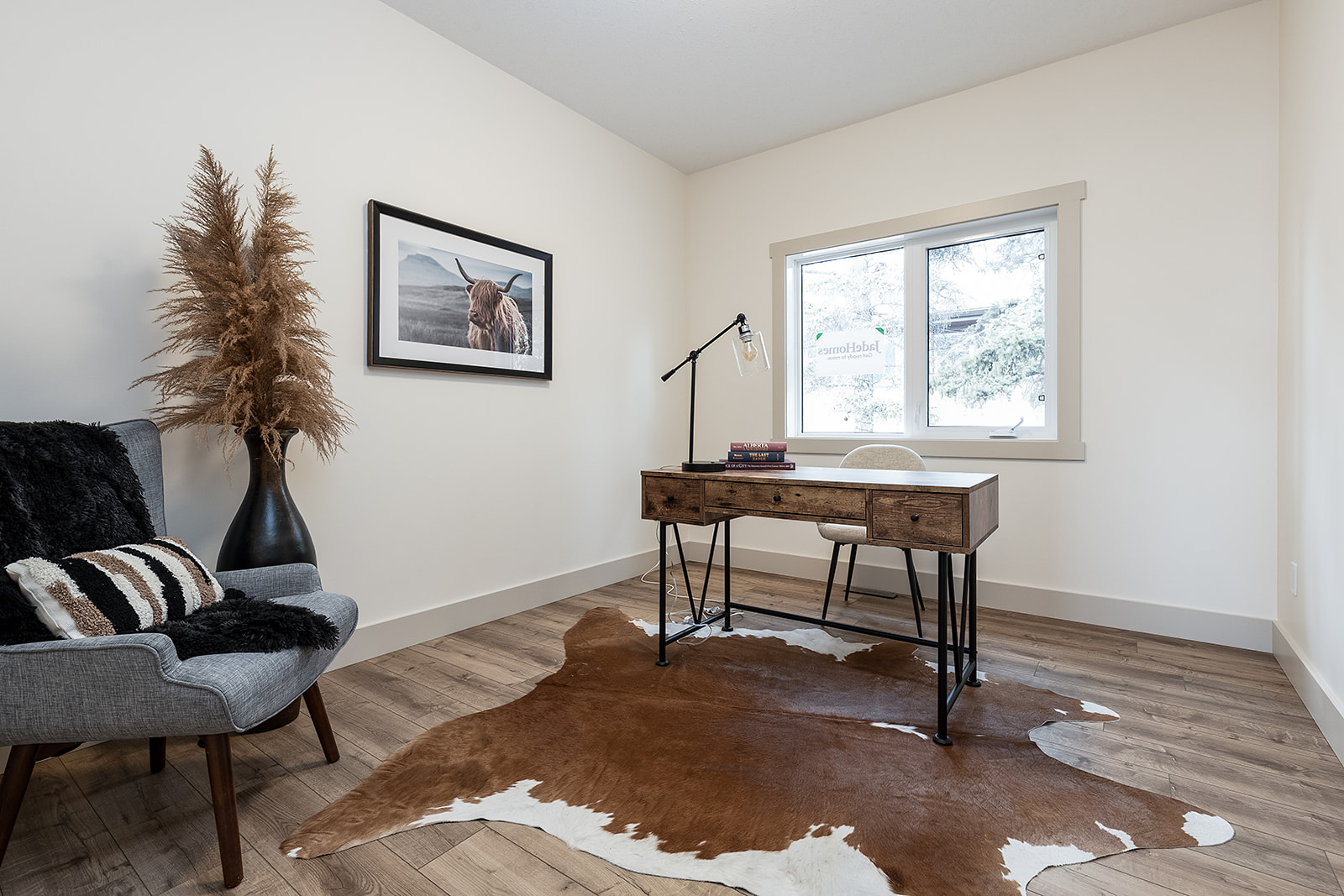 Modern office with a wooden desk on a cowhide rug, a framed highland cow photo, pampas grass in a vase, and a grey chair, creating a cozy ambiance.