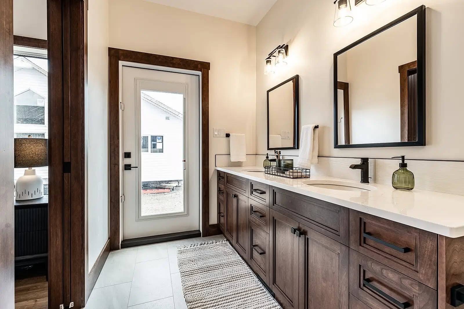 Modern bathroom with dual sinks and large mirrors framed in dark wood. Natural light from door, patterned rug on tile floor, and elegant, serene ambiance.
