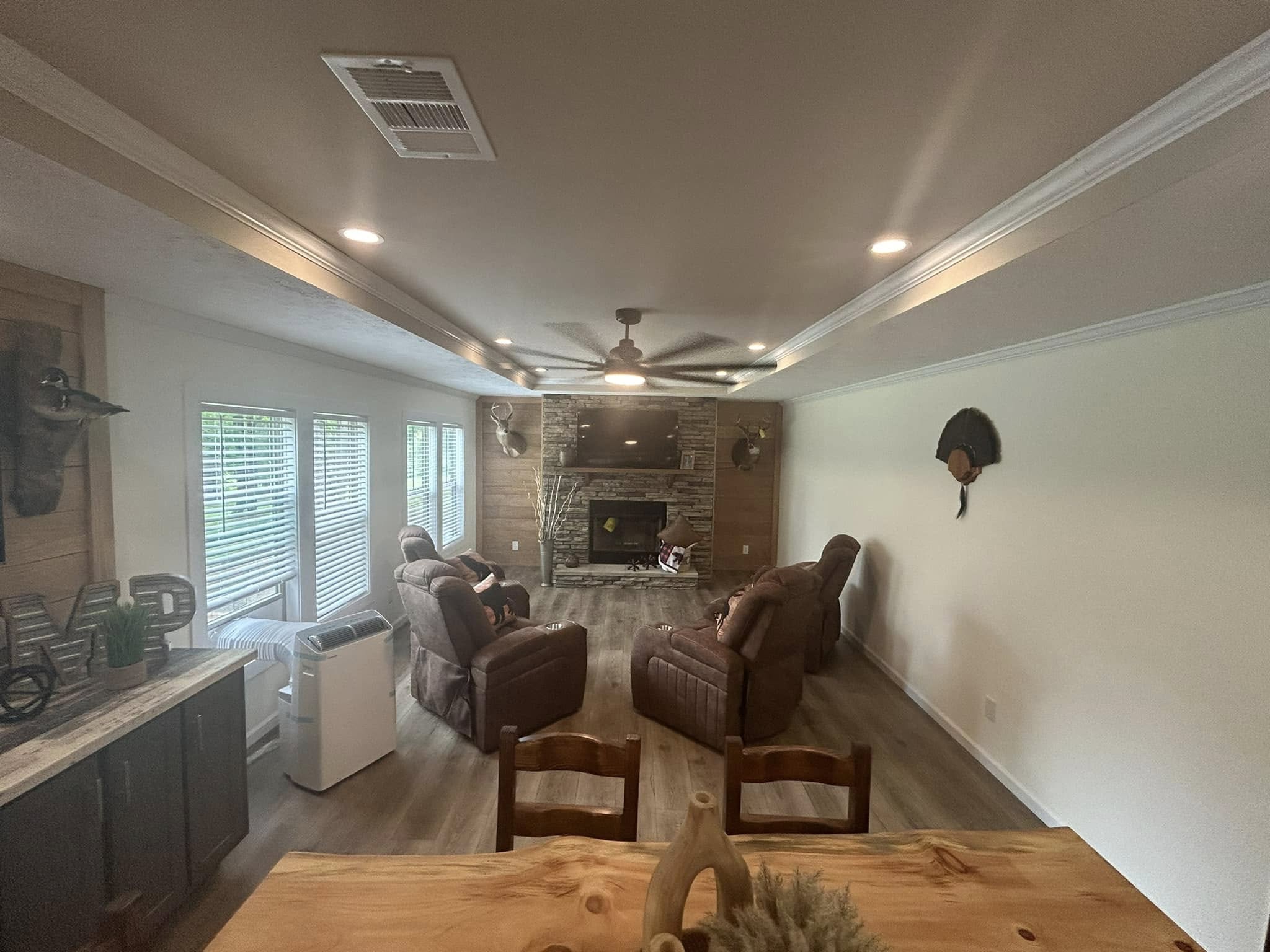 Cozy living room with two brown recliners facing a stone fireplace and mounted TV. Wooden decor and plants add warmth. Large windows on the left.