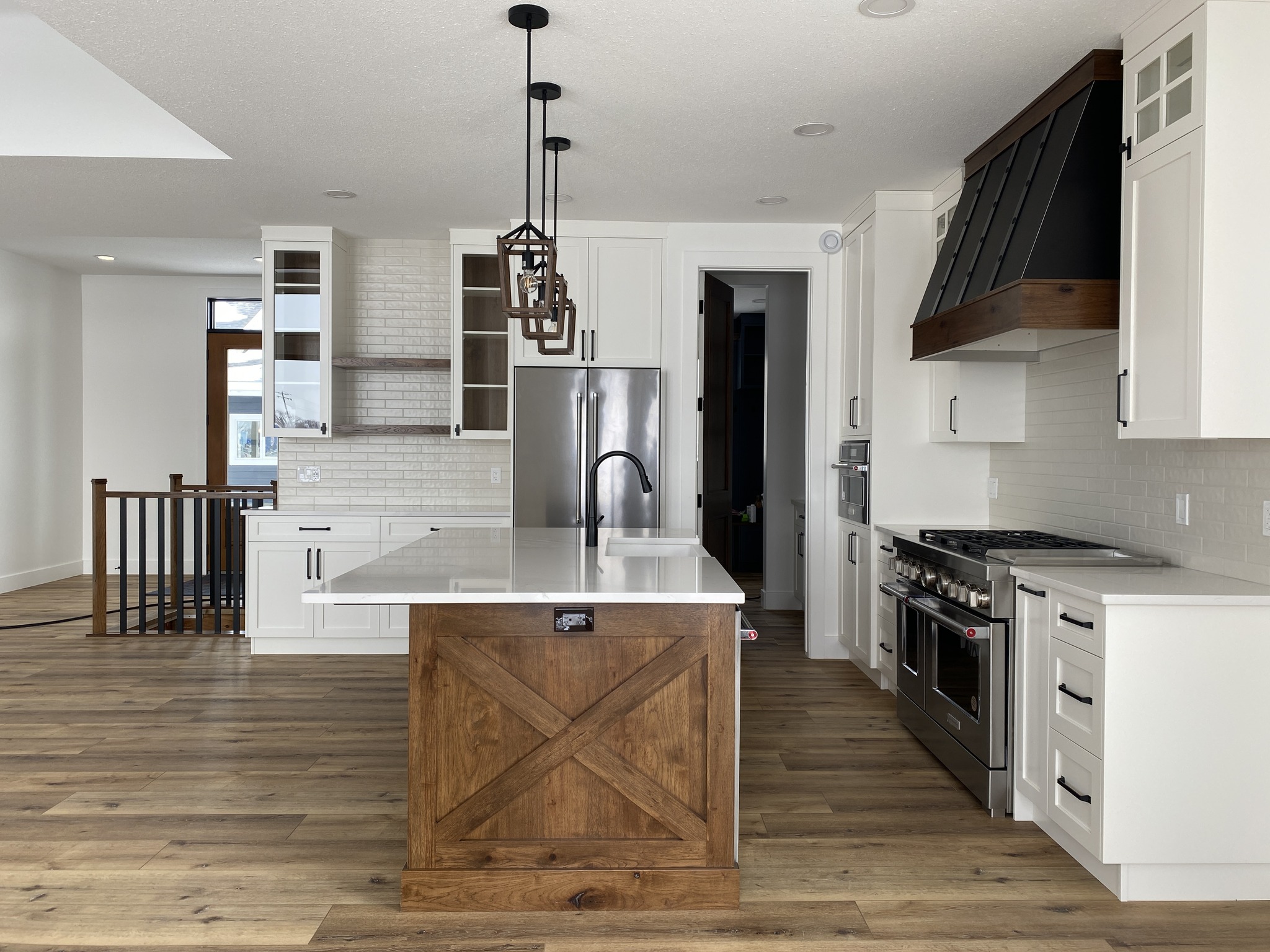 Spacious modern kitchen with a bright white island, wooden base, and hanging lights. White cabinets, stainless steel fridge, and stove. Warm, inviting tone.