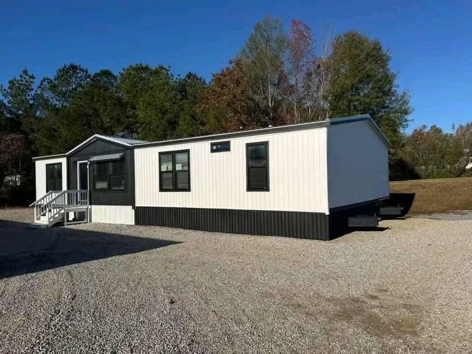 A white mobile home with black trim sits on a gravel lot, surrounded by tall trees under a clear blue sky, evoking a peaceful, rural feel.
