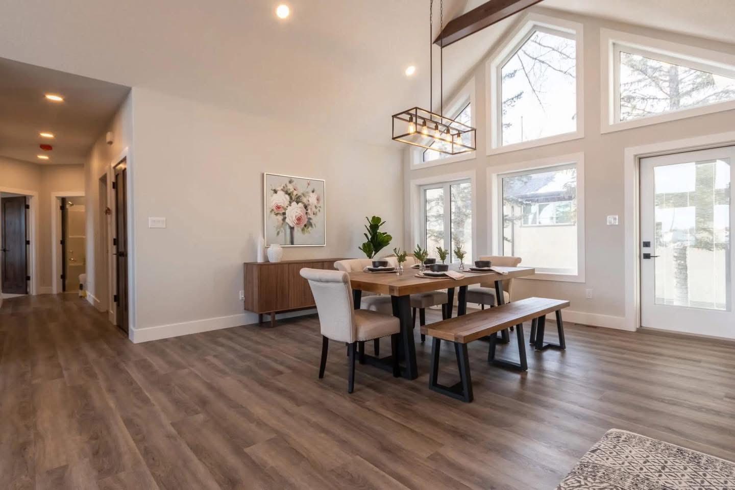 Bright dining room with vaulted ceilings, large windows, and wood flooring. Features a wooden table with benches, modern chandelier, and wall art. Cozy and inviting.